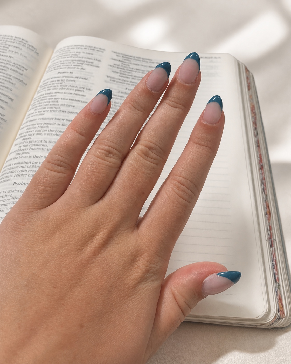 Long almond nails with deep teal French tips resting over an open book